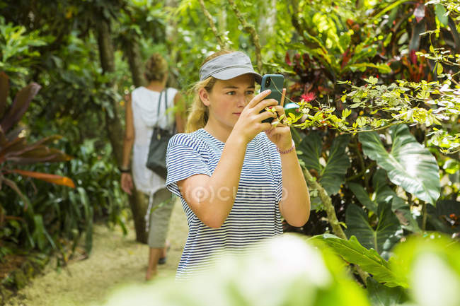 Adolescente rubia fotografiando follaje tropical verde con teléfono inteligente en el sendero de la naturaleza . - foto de stock