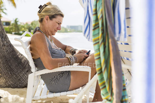 Ejecutiva femenina adulta usando smartphone en la playa, Isla Gran Caimán - foto de stock