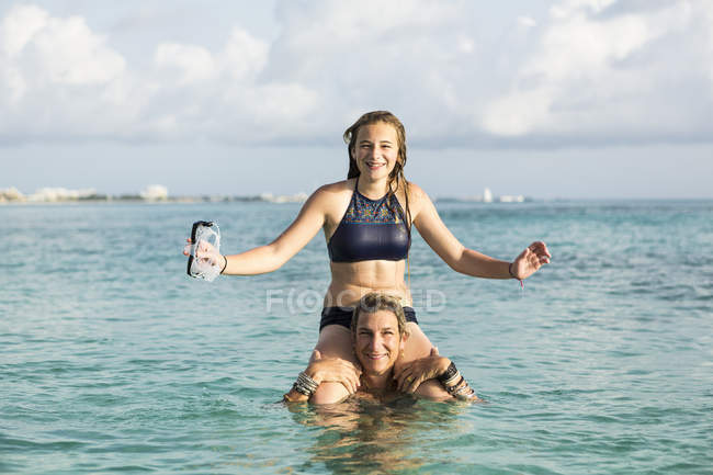 Adult woman standing in ocean water at sunset with daughter on shoulders — Stock Photo