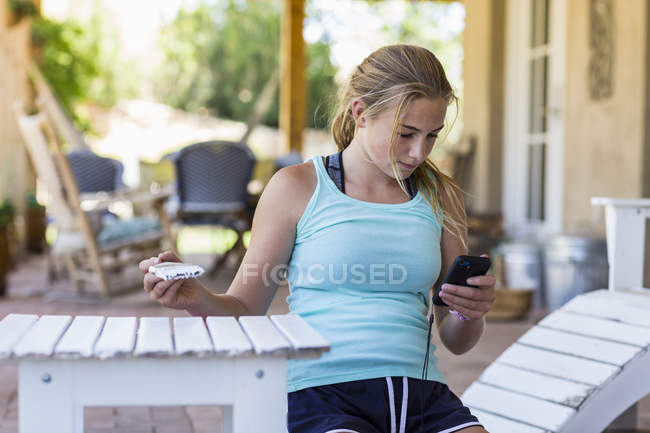 Blondes Teenie-Mädchen benutzt Smartphone beim Bemalen von Gartenmöbeln in Weiß. — Stockfoto
