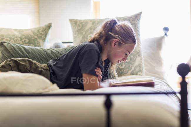 Teenage girl lying in bed and reading by window light. — Stock Photo