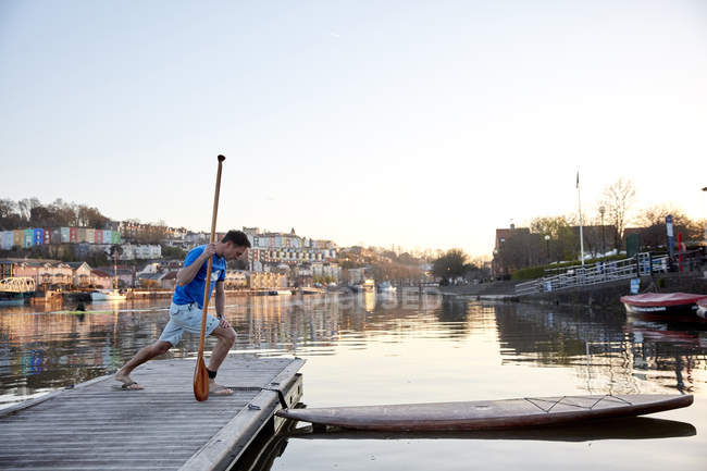 Homme tenant la pagaie étirant les jambes sur la jetée au bord de la rivière avant d'utiliser la planche à pagaie — Photo de stock