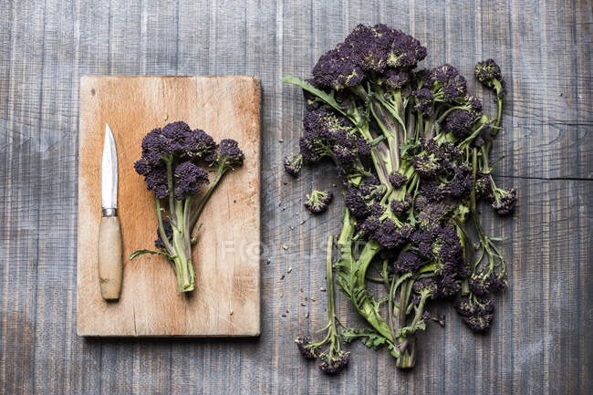 High angle close-up of purple sprouting broccoli, knife and wooden cutting board. — Stock Photo