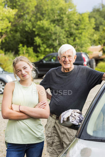 Senior grandfather and teenage granddaughter posing while washing car together in driveway — Stock Photo
