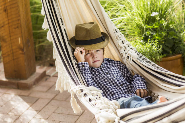 Cute elementary age boy in hat lying in hammock on porch — Stock Photo