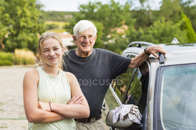 Abuelo mayor y nieta adolescente posando mientras lavan el coche juntos en la entrada - foto de stock
