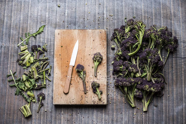 High angle close-up of purple sprouting broccoli, knife and wooden cutting board. — Stock Photo