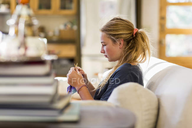 Vista laterale della ragazza adolescente che lavora a maglia sul divano in soggiorno — Foto stock