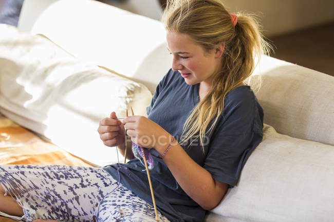 High angle view of smiling teenage girl with ponytail knitting on couch in early morning light — Stock Photo