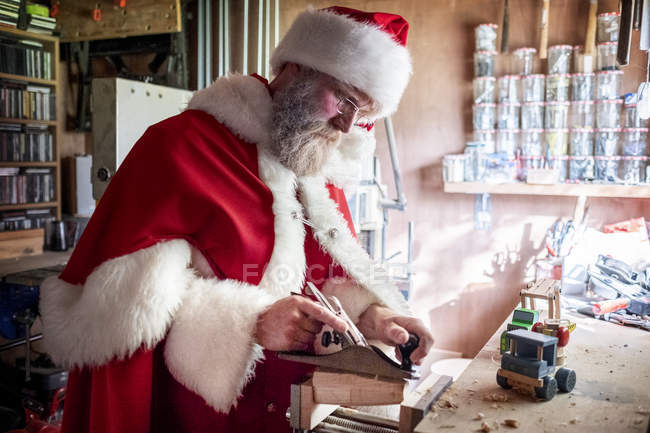 Man wearing Santa Claus costume standing in workshop and building wooden toy car. — Stock Photo