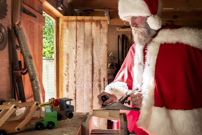 Uomo che indossa il costume di Babbo Natale in piedi in officina e costruzione di auto giocattolo in legno . — Foto stock