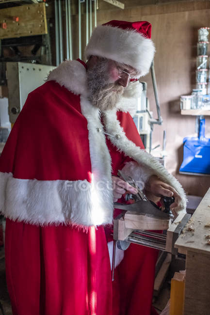 Homem vestindo traje de Papai Noel de pé em uma oficina e usando avião de madeira . — Fotografia de Stock