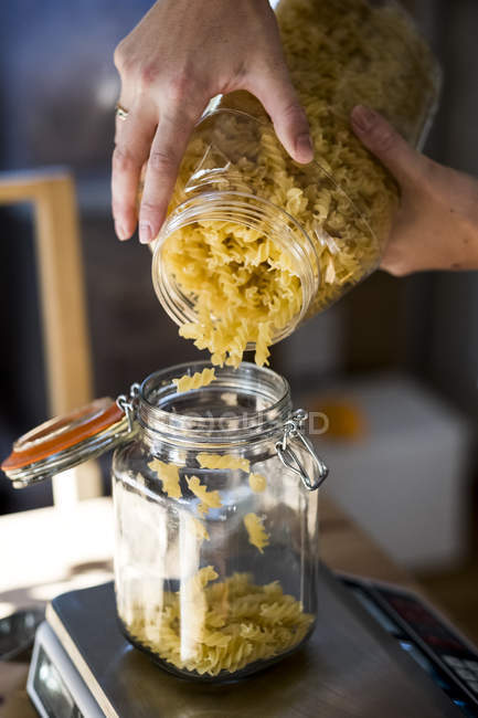 Крупный план человека, заливающего макароны Фузилли в стеклянную банку на кухонных весах . — стоковое фото