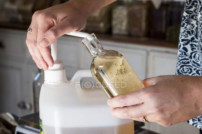 Mains de la personne debout dans une cuisine, décanter le liquide du récipient en plastique dans une bouteille en verre
. — Photo de stock
