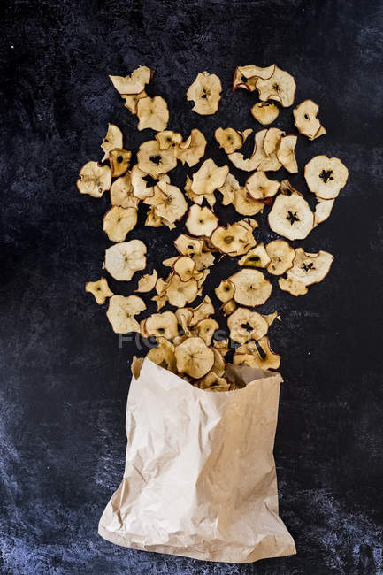 High angle close-up of paper bag and dried slices of apple on dark background. — Stock Photo