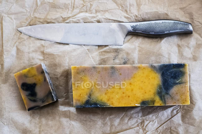 High angle close-up of yellow and black homemade bar of soap and kitchen knife. — Stock Photo