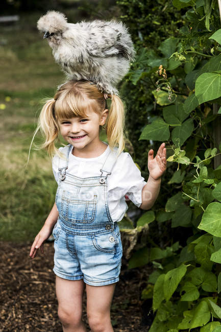 Lächelndes blondes Mädchen, das im Garten steht, mit flauschigem grauen Huhn auf dem Kopf. — Stockfoto
