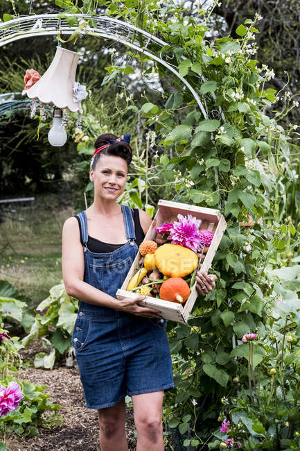 Woman standing in garden, holding wooden crate with fresh vegetables and flowers, smiling in camera. — Stock Photo