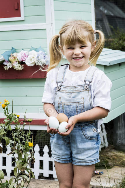 Blonde girl standing in garden in front of hen house, holding fresh eggs. — Stock Photo