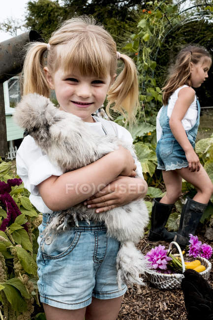 Blonde girl standing in garden, holding fluffy grey chicken. — Stock Photo