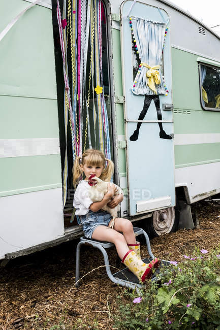 Blonde girl holding white chicken sitting outside a caravan. — Stock Photo
