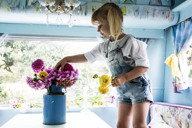 Blonde girl standing at table inside blue caravan, putting pink Dahlias into blue vase. — Stock Photo
