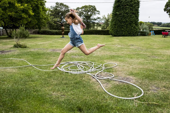 Mädchen in Jeans-Latzhose springt über Gartenschlauch auf grünen Rasen. — Stockfoto