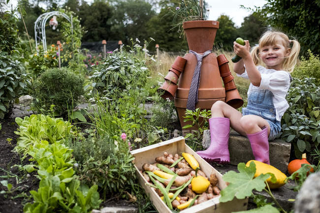 Smiling blonde girl sitting in garden next to terracotta scarecrow, picking fresh vegetables. — Stock Photo