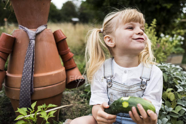 Blonde girl sitting in garden next to terracotta scarecrow, eating cucumber. — Stock Photo