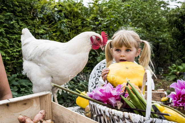 Blonde girl holding yellow gourd and white chicken in garden. — Stock Photo