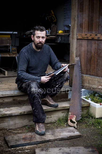 Bearded man sitting on steps outside workshop, holding handmade knife, looking in camera. — Stock Photo
