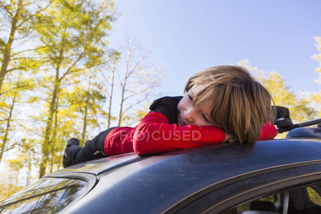 Sonriente niño pre-adolescente acostado en la capucha de SUV azul en el bosque . - foto de stock