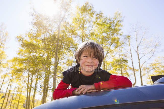 Elementary age boy lying on hood of blue SUV in woodland. — Stock Photo
