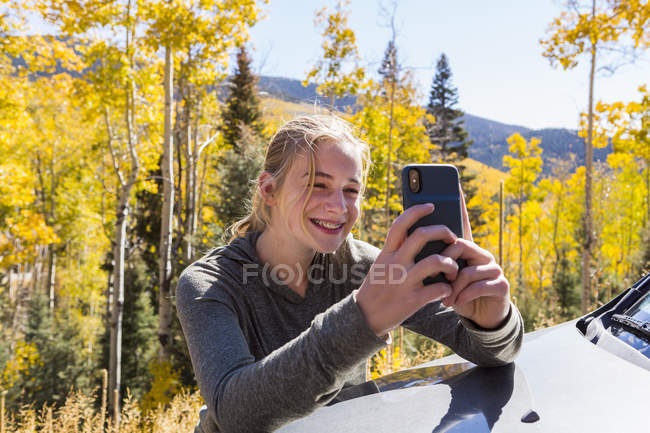 Chica adolescente tomando fotos con teléfono inteligente, mirando los árboles de álamo de otoño - foto de stock
