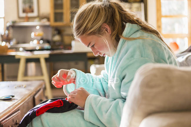 Adolescente coser uma camisa sentada em roupão de banho em casa . — Fotografia de Stock