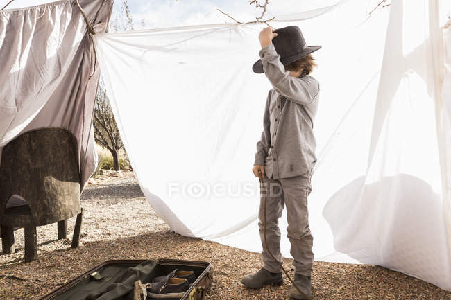 Menino da idade elementar usando chapéu jogando na tenda ao ar livre feita de lençóis — Fotografia de Stock