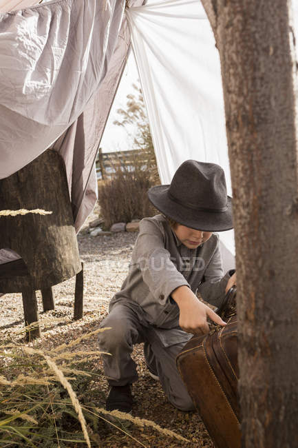 Menino de idade elementar usando chapéu brincando com mala em tenda ao ar livre feita de lençóis — Fotografia de Stock
