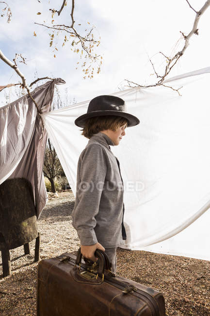 Elementary age boy wearing hat carrying luggage in outdoor tent made of sheets — Stock Photo