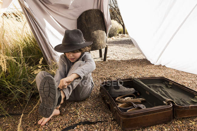 Menino da idade elementar usando chapéu colocando bota na tenda ao ar livre feita de lençóis — Fotografia de Stock
