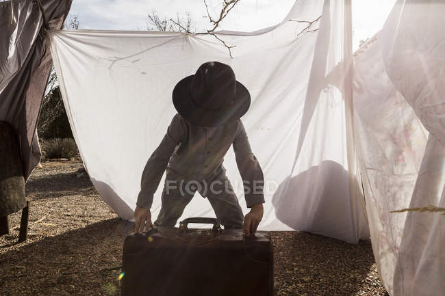 Мальчик младшего возраста в шляпе играет с чемоданом в наружной палатке из простыней — стоковое фото