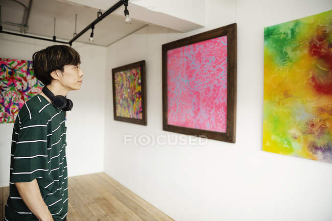 Japanese man wearing headphones looking at abstract paintings in an art gallery. — Stock Photo