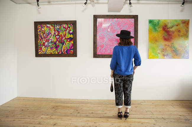 Rückansicht einer Frau mit Hut, die vor abstrakten Gemälden in einer Kunstgalerie steht. — Stockfoto