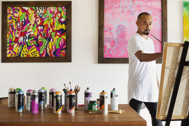 Japanese man standing in art gallery, holding paintbrush, working on artwork on easel. — Stock Photo