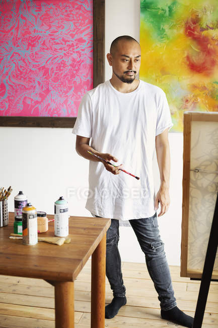 Japanese artist standing in art gallery, holding paintbrush, looking at artwork on easel. — Stock Photo