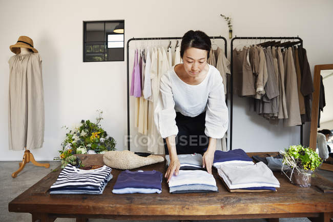 Femme japonaise debout dans une petite boutique de mode, arrangeant des t-shirts sur une table basse . — Photo de stock