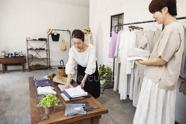 Zwei japanische Frauen stehen in einer kleinen Modeboutique und arrangieren T-Shirts auf einem Couchtisch. — Stockfoto