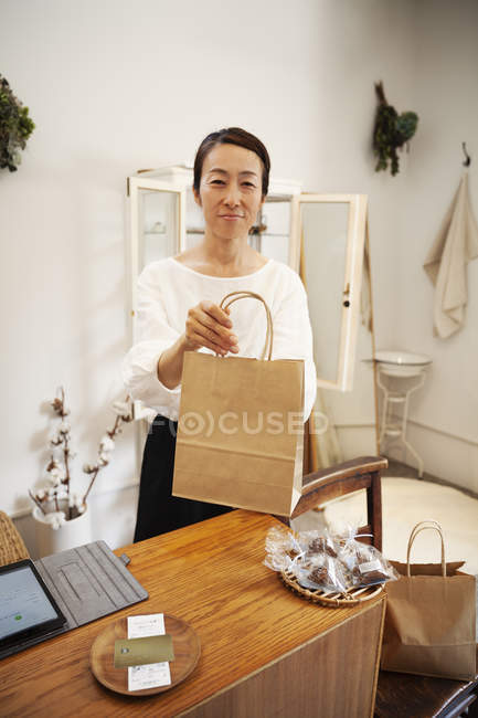 Посміхаючись, японська жінка стоїть у маленькому бутіку, тримаючи коричневий паперовий пакет, дивлячись у камеру.. — стокове фото