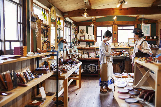 Japanese woman wearing apron standing in a leather shop, holding leather clutch bag, talking to customer. — стокове фото