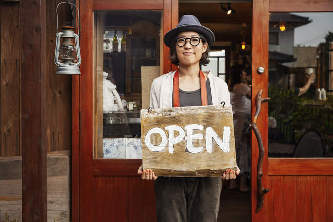 Mulher japonesa usando chapéu e óculos de pé na frente de uma loja de couro, segurando o sinal aberto . — Fotografia de Stock