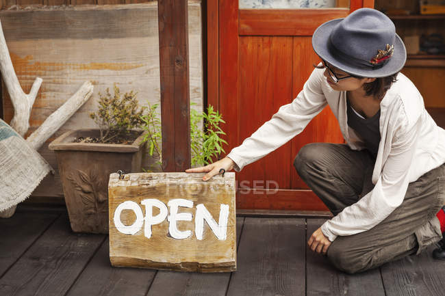 Japanese woman wearing hat and glasses kneeling in front of a leather shop, holding Open sign. — Stock Photo